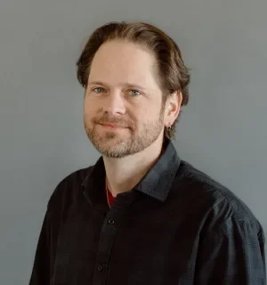 Chris Albrecht wearing a dark button down shirt in front of a gray background.