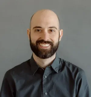 Chris DeLuca wearing a dark button down shirt in front of a gray background.