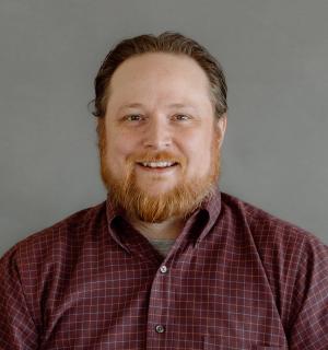 David Burns wearing a red check button down shirt in front of a gray background.
