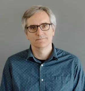 Greg Dunlap wearing a teal button down shirt in front of a gray background.