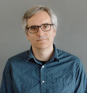 Greg Dunlap wearing a teal button down shirt in front of a gray background.