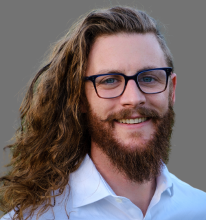 Hawkeye Tenderwolf wearing a blue button down shirt and glasses in front of a gray background.
