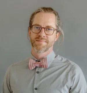 James Sansbury wearing a gray button down shirt with a pink and white striped bowtie in front of a gray background.