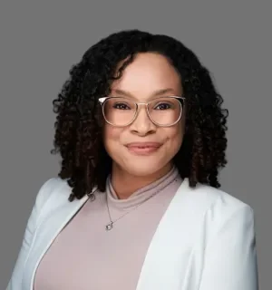 Jordan Gonzalez wearing glasses, a white blazer, and a pink blouse in front of a gray background.