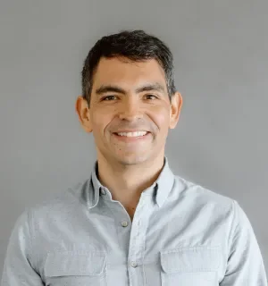 Juampy NR wearing a blue button down shirt standing in front of a gray background.