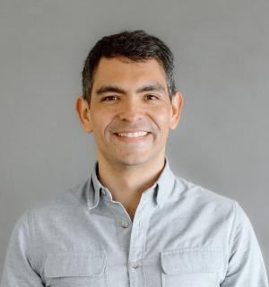 Juampy NR wearing a blue button down shirt standing in front of a gray background.