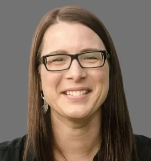 Kris Konrady wearing glasses and wearing a black button down shirt in front of a gray background.