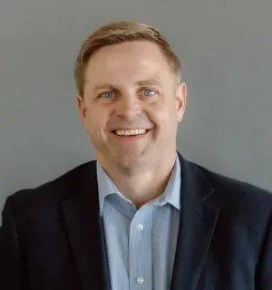 Matt Kleve wearing a blue button down shirt with a navy sport coat in front of a gray background.