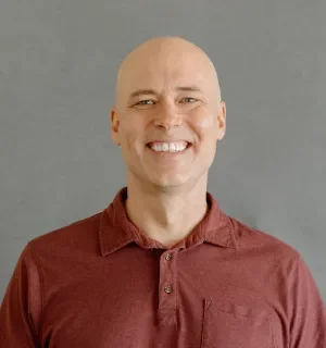 Matthew Tift wearing a red button down shirt in front of a gray background.