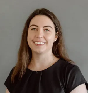 Megh Plunkett wearing a black short-sleeved shirt in front of a gray background.