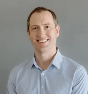 Nate Lampton wearing a blue biutton down shirt in front of a gray background.