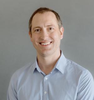 Nate Lampton wearing a blue biutton down shirt in front of a gray background.