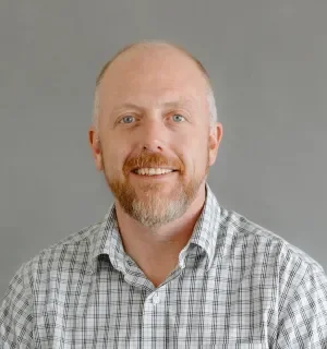 Ned Lucks wearing a white button down shirt with black plaid overlay in front of a gray background.