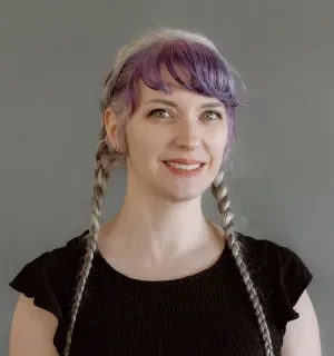 Pauline Judge wearing a short-sleeved black shirt and braided pigtails with purple hair in front of a gray background.