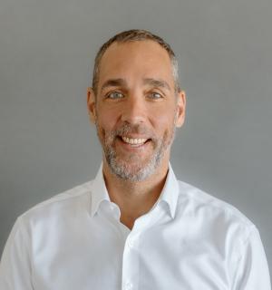 Photo of Seth Brown, white male wearing a white button down oxford shirt in front of a gray background.