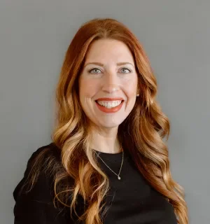 Stephanie Ganzer wearing a black long-sleeved t-shirt and gold necklace in front of a gray background.