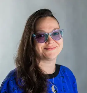 Marissa Epstein is a woman in her mid-30s with long brown hair shaved on one side, wearing a purple and black blouse, purple cat-eye glasses, a silver necklace, and a smirk.