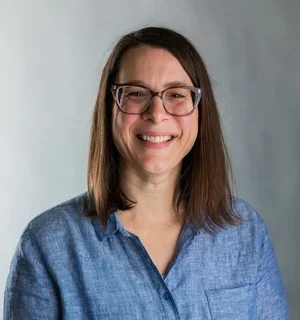 A smiling woman with straight brown hair and glasses, wearing a light blue button-up shirt, standing in front of a neutral gray background.