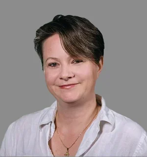 Professional headshot of a woman with short, side-swept brown hair and fair skin, smiling gently at the camera. She is wearing a white button-down shirt and a delicate gold necklace, posed against a plain gray background.