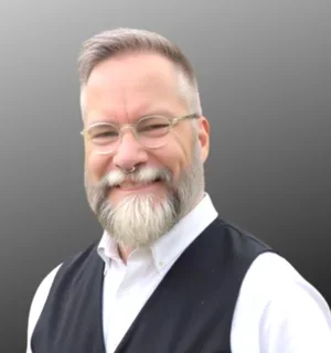 Portrait of a smiling man with short gray hair, a full gray beard, and round glasses, wearing a white button-down shirt and dark vest against a neutral gray background.