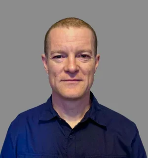 A person with a closely cropped haircut and small hoop earrings stands facing the camera against a plain light-gray wall, wearing a dark navy button-up shirt and a slight smile.