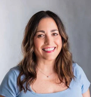 Woman with long, wavy brown hair, brown eyes, wearing a light blue scoop-neck t-shirt in front of a gray background.