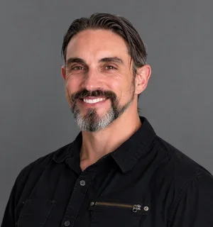 Smiling man with dark, graying hair and a salt-and-pepper beard, wearing a black button-up shirt against a medium-gray background.