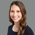 Headshot of white woman with brown hair, smiling, wearing a black shirt in front of a grey background.