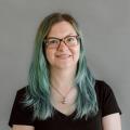 Aubrey Sambor with green hair wearing a black t-shirt in front of a gray background.