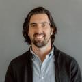 Brian Skowron wearing blue button down shirt with dark gray sweater in front of a gray background.