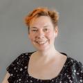 Cathy Theys with orange hair wearing a black blouse with white flowers in front of a gray background.