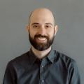 Chris DeLuca wearing a dark button down shirt in front of a gray background.