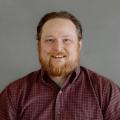 David Burns wearing a red check button down shirt in front of a gray background.