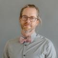 James Sansbury wearing a gray button down shirt with a pink and white striped bowtie in front of a gray background.