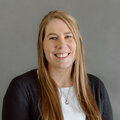 Kayla AhoWalsh wearing a black sweater with white t-shirt in front of a gray background.