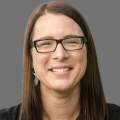 Kris Konrady wearing glasses and wearing a black button down shirt in front of a gray background.