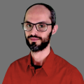 Marcos Cano wearing a red button down shirt and wearing glasses in front of a gray background.