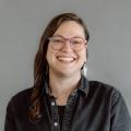 Marissa Epstein wearing glasses and a black button down shirt in front of a gray background.