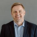 Matt Kleve wearing a blue button down shirt with a navy sport coat in front of a gray background.