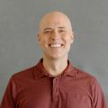 Matthew Tift wearing a red button down shirt in front of a gray background.