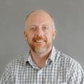 Ned Lucks wearing a white button down shirt with black plaid overlay in front of a gray background.