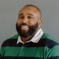 Riche Zamor wearing a blue and green striped rugby shirt with white collar in front of a gray background.