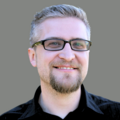 Headshot of Jerad Bitner wearing glasses and black shirt on gray background.