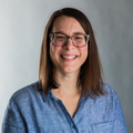 A smiling woman with straight brown hair and glasses, wearing a light blue button-up shirt, standing in front of a neutral gray background.
