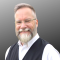 Portrait of a smiling man with short gray hair, a full gray beard, and round glasses, wearing a white button-down shirt and dark vest against a neutral gray background.